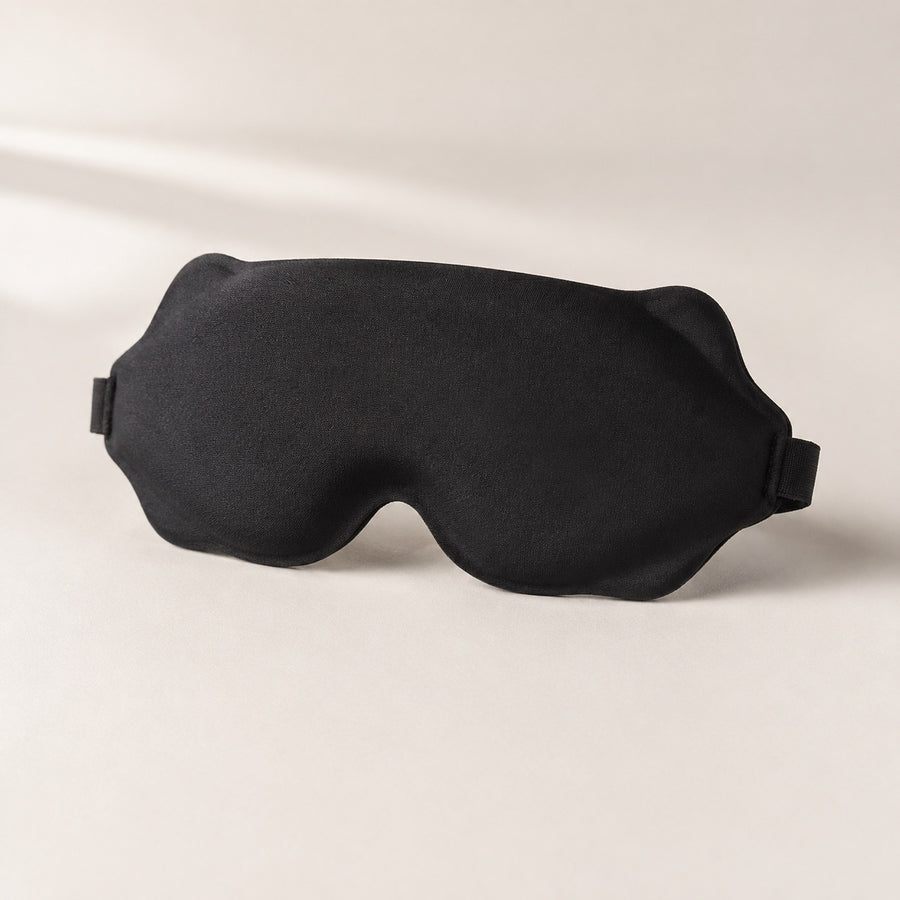 Black contoured sleep mask on a warm ivory background, showing the outer face.