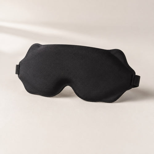 Black contoured sleep mask on a warm ivory background, showing the outer face.